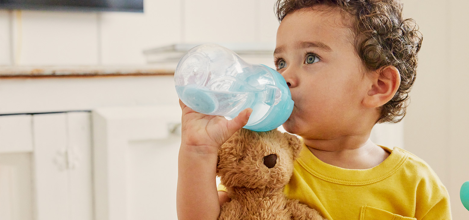 Toddler in yellow shirt drinking from a blue and clear sippy cup while holding a brown teddy bear.