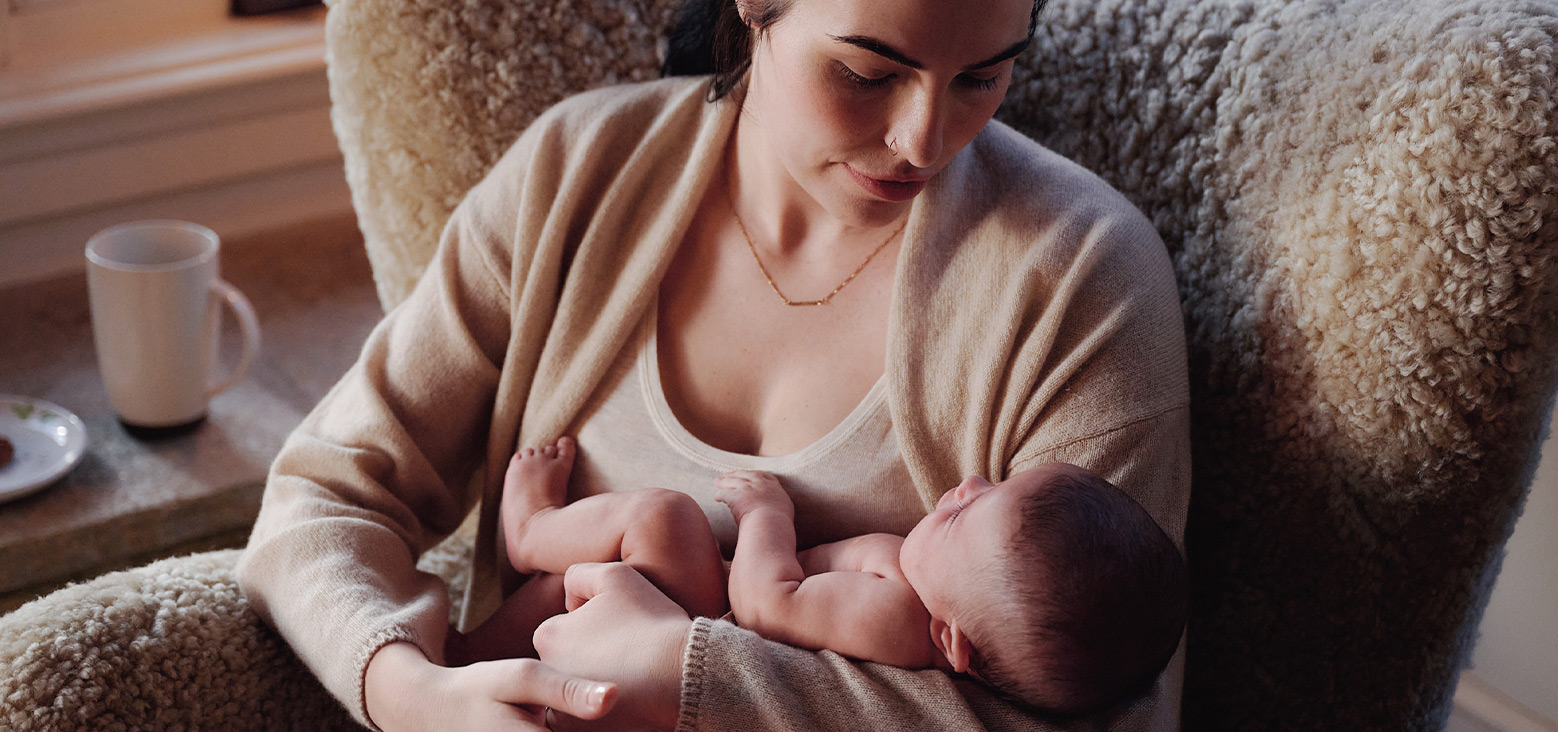 Mother holding newborn baby while sitting in chair, warm home setting with mug on side table
