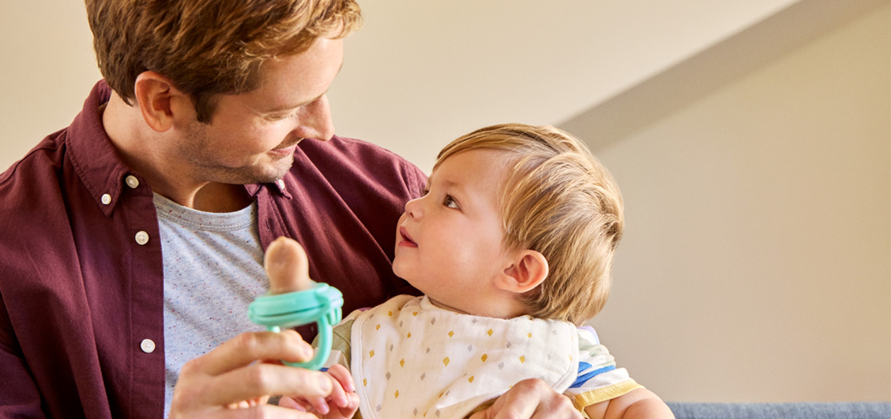 Father holding toddler who is playing with a turquoise teething ring toy.