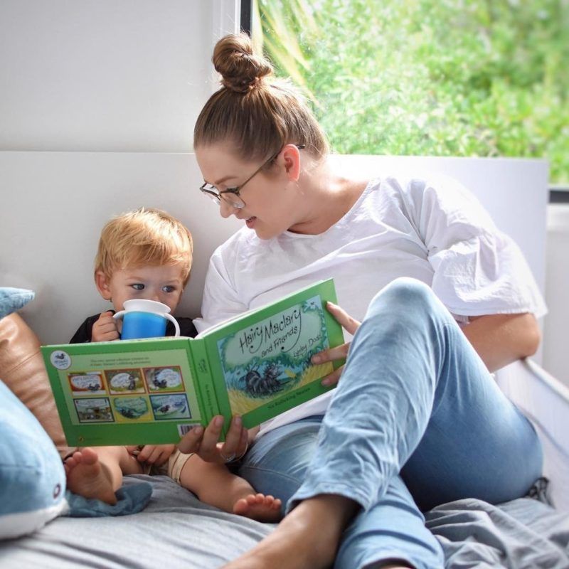 Adult and toddler reading a green book together while toddler drinks from a blue cup by a window.