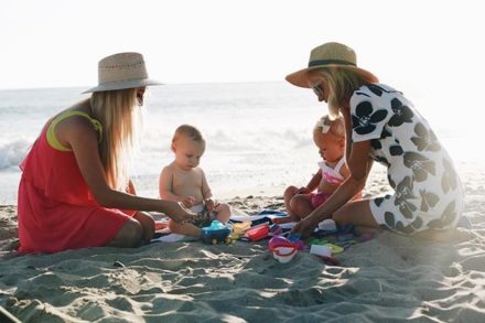 Two adults and two toddlers playing with colorful toys on a sandy beach near the ocean.
