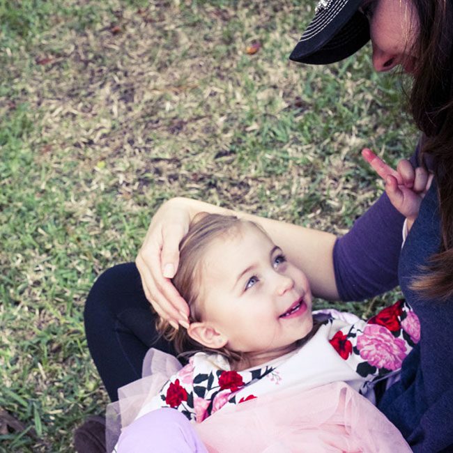 Toddler in a pink tutu and floral shirt looking up while sitting on grass with an adult nearby.