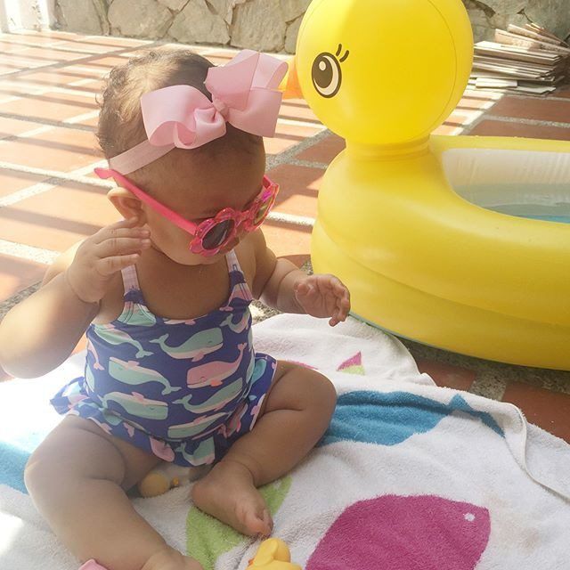 Toddler in a colorful swimsuit and pink bow headband sitting poolside next to a large yellow inflatable duck toy.