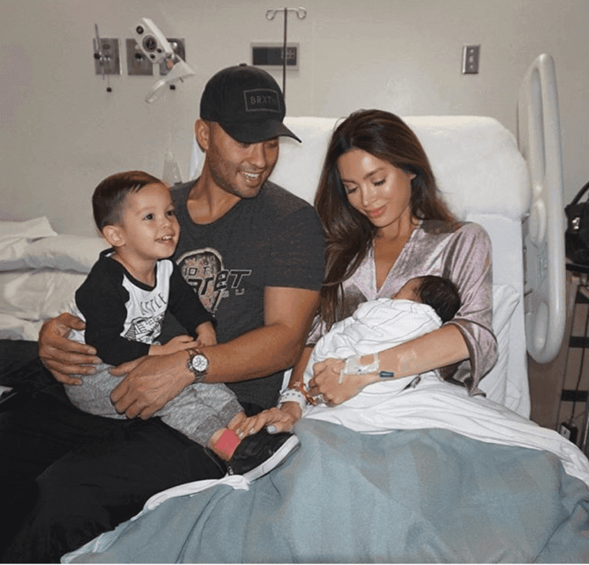 Family with two children in a hospital bed, parents smiling while holding newborn and toddler.
