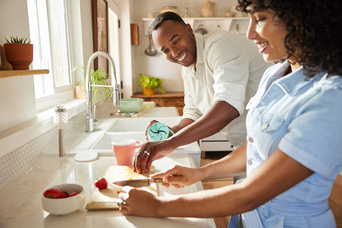 Parents preparing food together in a bright kitchen, man holding a Munchkin bowl while woman chops vegetables on a cutting board.