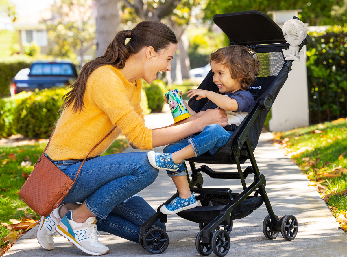 Woman giving a toddler a drink from a Munchkin sippy cup while sitting in a Munchkin stroller on a sunny residential street.