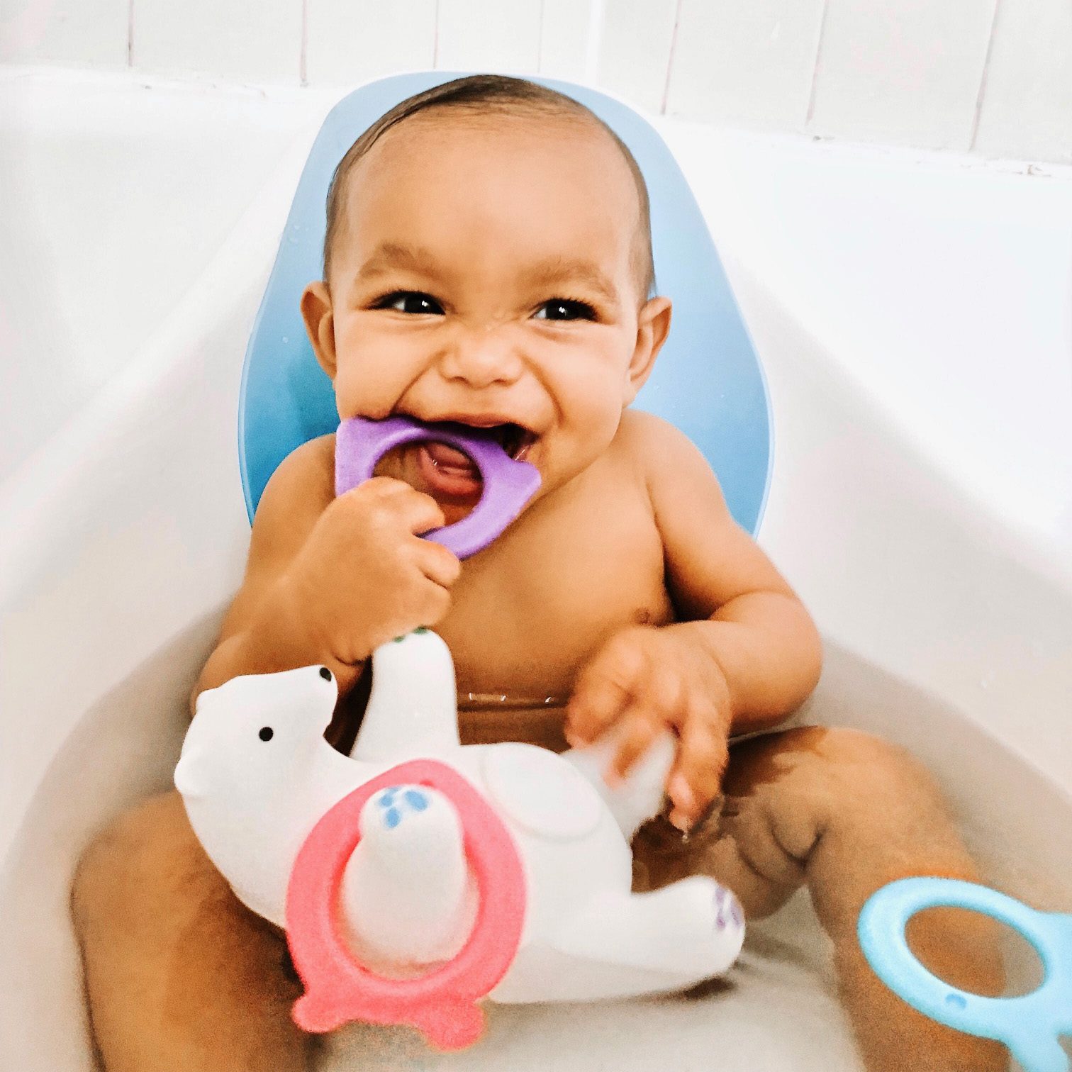 Baby chewing on a purple teething toy while holding a white duck-shaped toy with pink and blue accents in a bathtub.