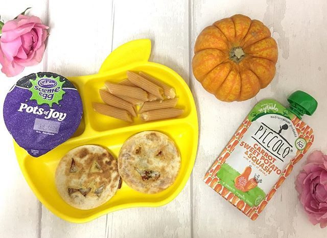 Yellow apple-shaped plate with two muffins and breadsticks, surrounded by Cadbury Pots of Joy yogurt, Piccolo food pouch, decorative pumpkin, and pink flowers on white wooden surface.
