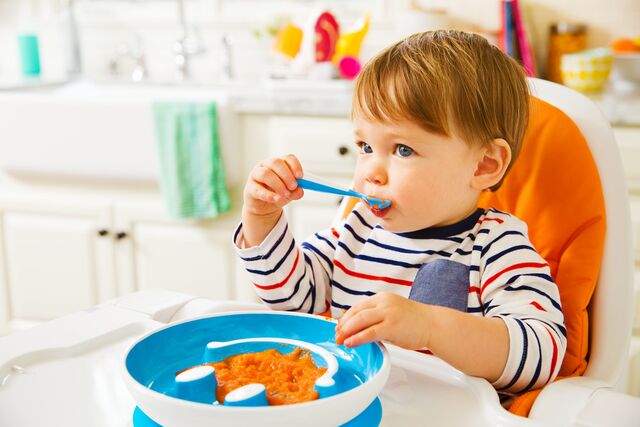 Toddler eating from a blue bowl with a spoon in a bright kitchen.