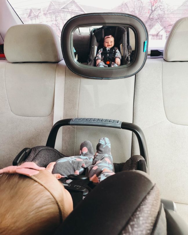 Munchkin car seat mirror showing toddler in rear-facing car seat, with parent's arm visible in foreground and cream-colored car interior.