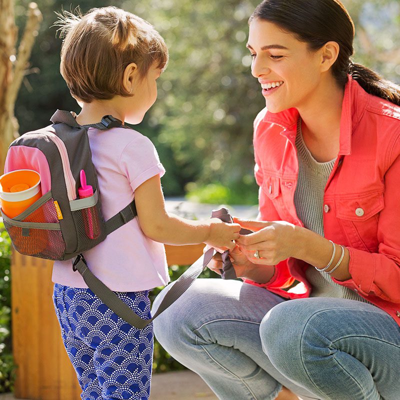 Woman and toddler outdoors, toddler wearing Munchkin backpack with orange and black design and mesh pockets, woman adjusting backpack straps while smiling at child.
