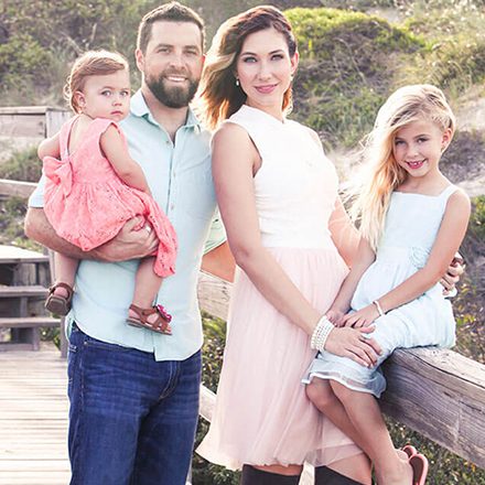 Family of four on a wooden deck with nature background, parents with two young daughters in casual summer clothing.