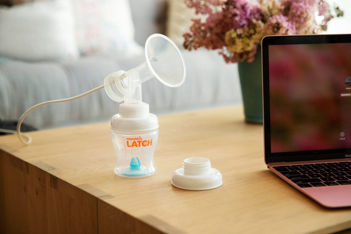 Munchkin Latch breast pump displayed on a wooden desk next to a laptop, with dried flowers and home decor in the background.