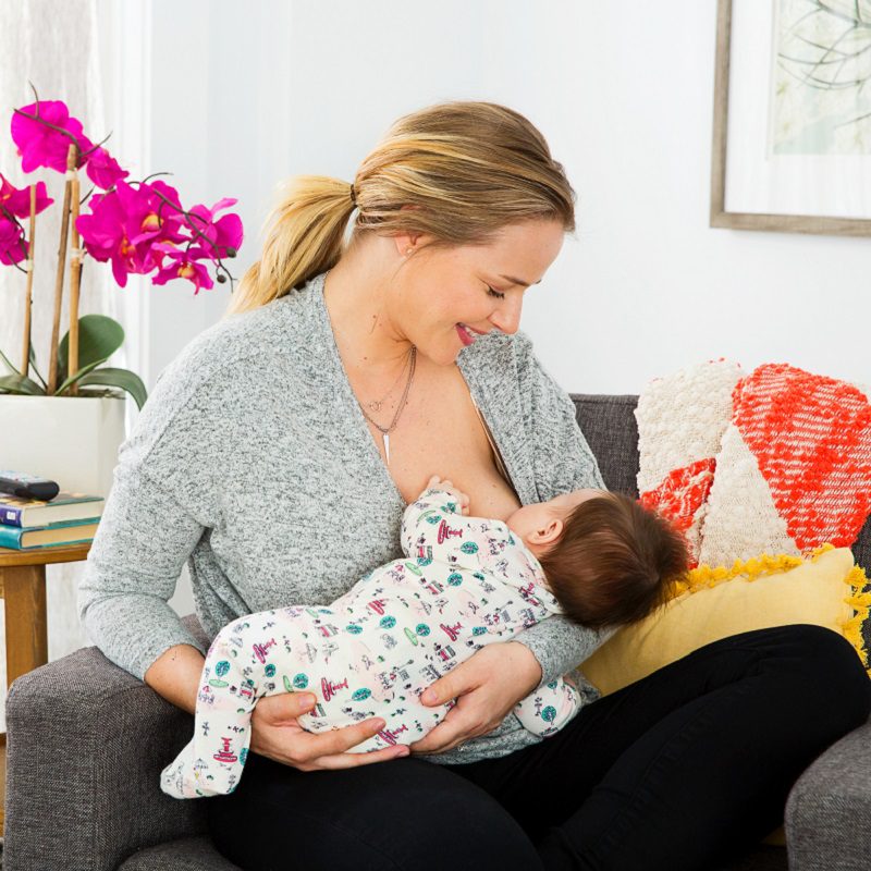 Woman breastfeeding infant on couch in bright living room with colorful blankets and pink flowers nearby