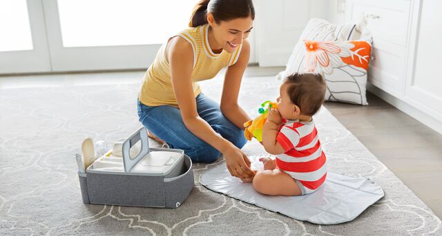Woman and toddler playing together on a mat, with a gray storage bin and colorful toys nearby in a bright room.