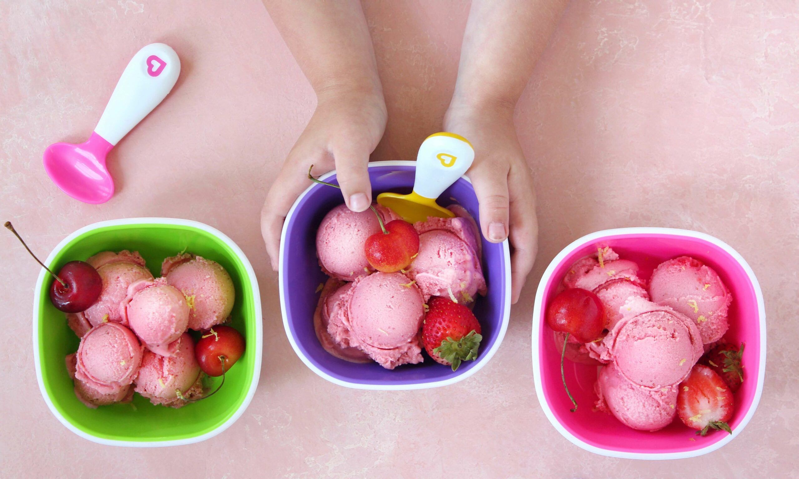 Child's hands scooping pink ice cream with Munchkin spoons from three colorful bowls filled with strawberry ice cream and fresh berries.