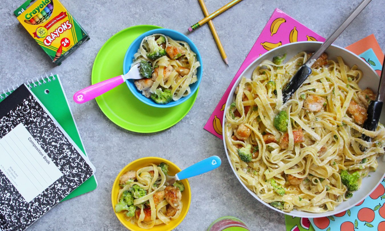 Overhead view of toddler meal setup with pasta and vegetables served in Munchkin bowls and plates in bright colors, alongside crayons, notebook, and utensils on a gray surface.