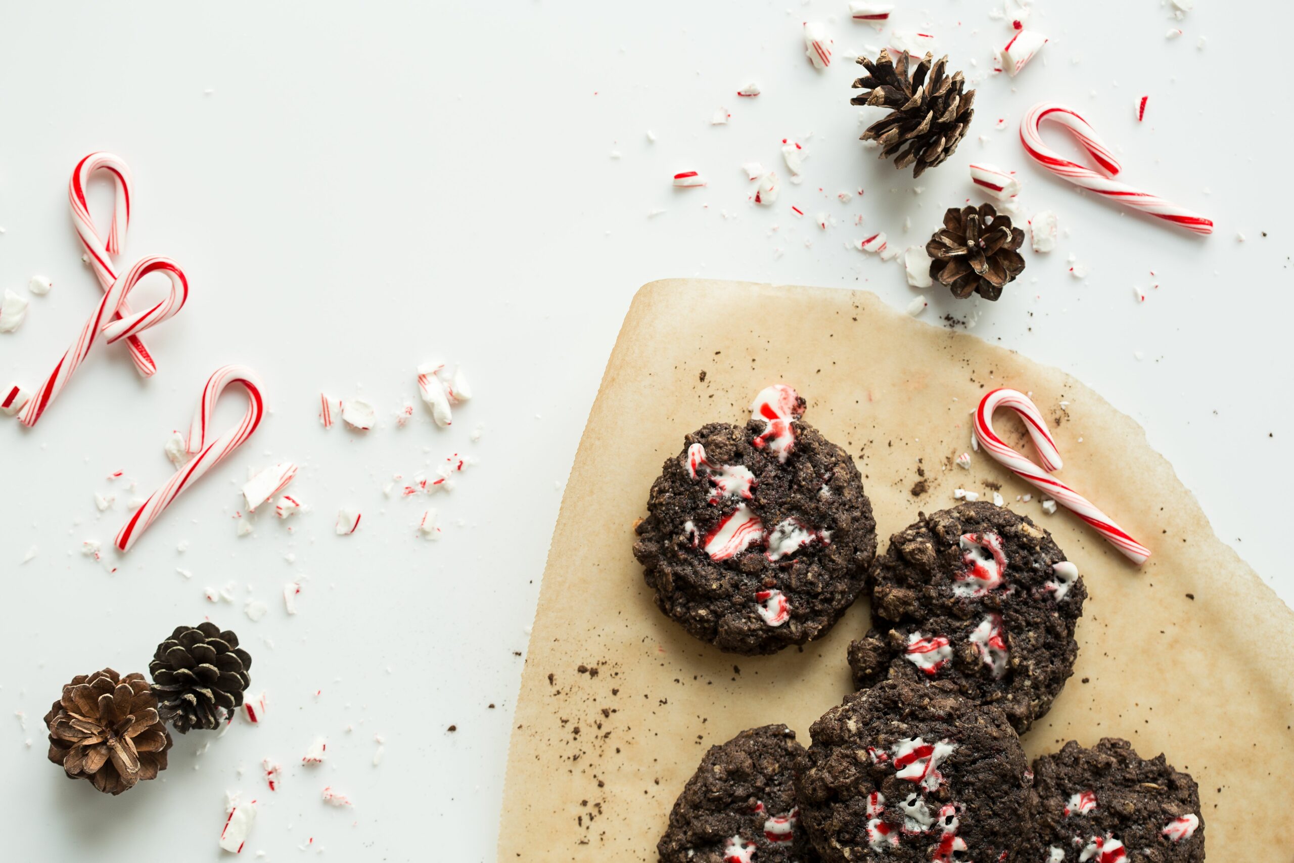 Chocolate cookies with crushed candy canes on parchment paper, surrounded by candy canes, pinecones, and peppermint pieces on a white surface.