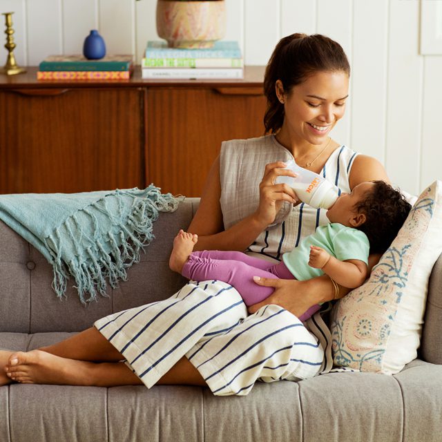 Woman feeding a toddler from a Munchkin sippy cup while sitting on a couch in a bright living room.