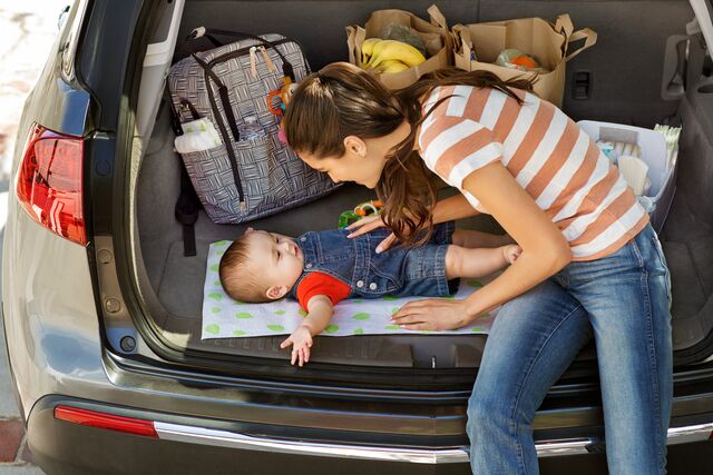 Parent and baby in open car trunk with groceries, bags, and Munchkin products organized for travel.