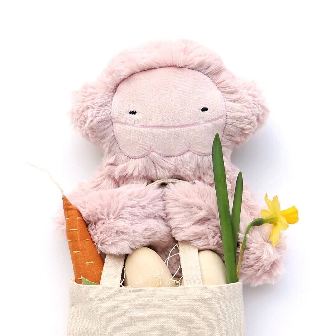 Pink plush bunny toy in a cream fabric pouch with carrot, eggs, daffodil flower, and green plant stems.
