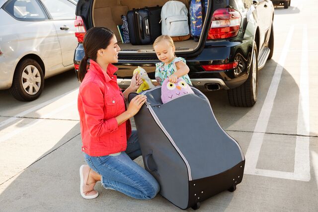 Woman kneeling beside a toddler sitting in a Munchkin car seat in a parking lot with an open vehicle trunk in the background.