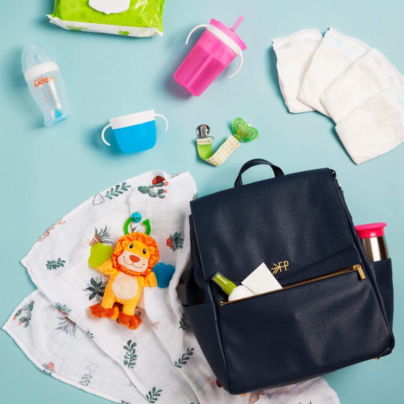 Flat lay of Munchkin baby products on light blue background including clear bottle, pink and blue sippy cups, white cloths, lion plush toy, teething toy, wipes package, and black diaper bag with contents visible.