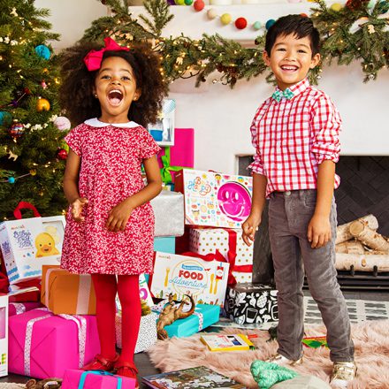 Two children smiling in front of a decorated Christmas tree surrounded by colorful wrapped gifts and toys.
