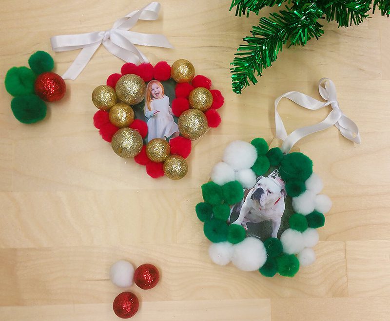 Two festive holiday ornaments with white ribbon loops displayed on a light wood surface: one features a baby photo surrounded by red and gold pom-poms and glitter balls, the other shows a dog photo surrounded by green and white pom-poms, with evergreen garland and loose ornaments nearby.