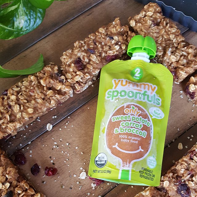 Yummy Spoonfuls sweet potato carrot broccoli baby food pouch on a wooden surface next to homemade granola bars with oats and dried fruit.