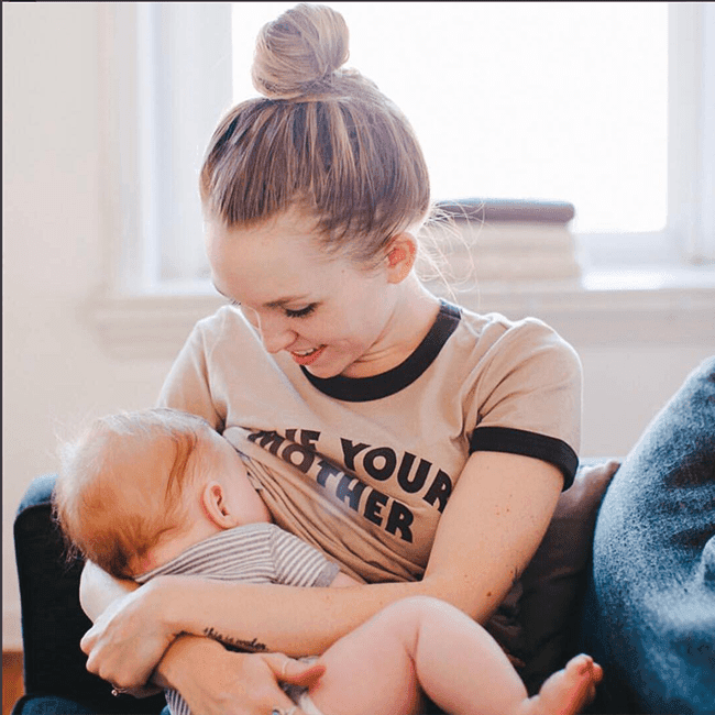Parent holding smiling infant in a bright room, parent wearing tan and black shirt with text.