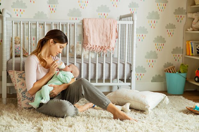 Woman sitting on a nursery floor holding and feeding a baby in a green blanket, with a white crib and cloud-patterned wallpaper in the background.
