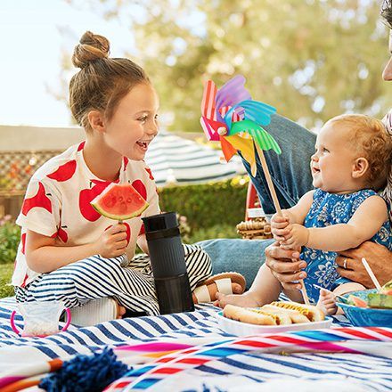 Older girl and toddler playing outdoors with a colorful pinwheel toy during a picnic, girl holding a watermelon slice.
