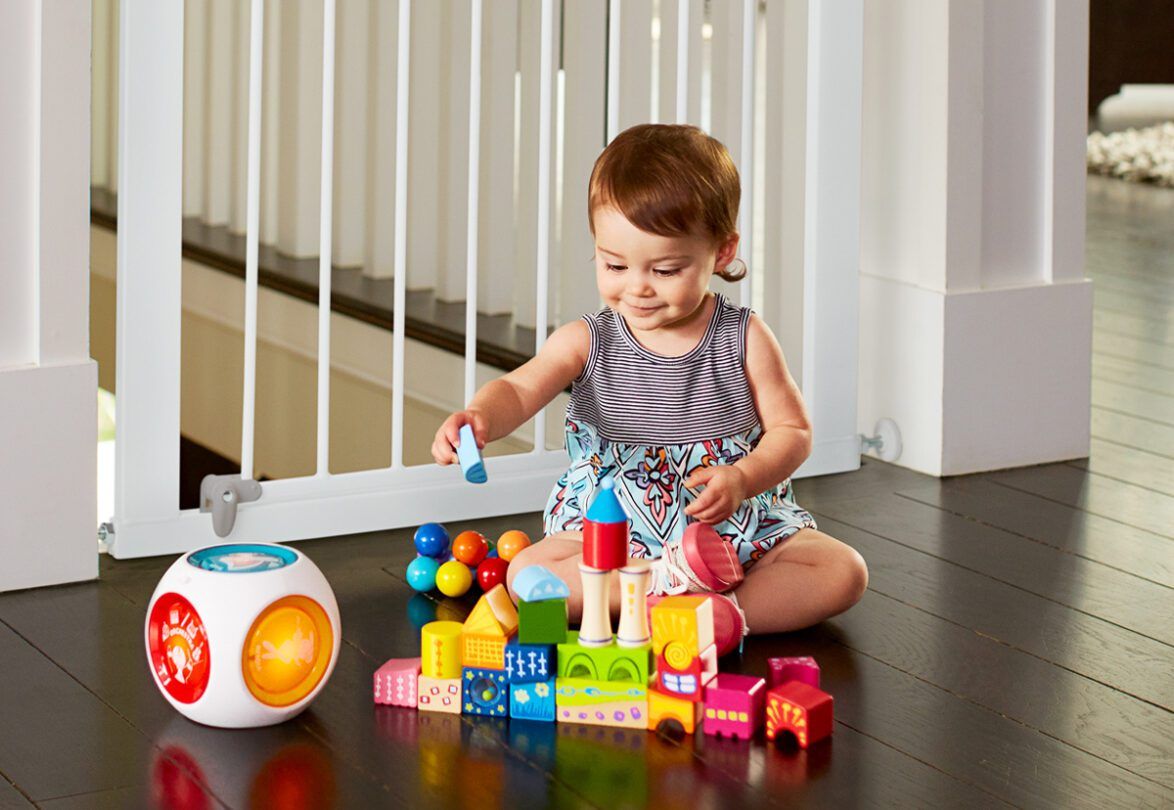 Toddler playing with colorful building blocks and a light-up activity toy on a dark floor in a playpen.