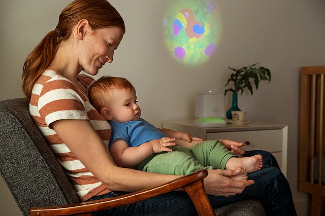 Woman holding a baby in her lap while watching a colorful light projection on the wall, with a potted plant and nightlight visible on a nearby shelf.