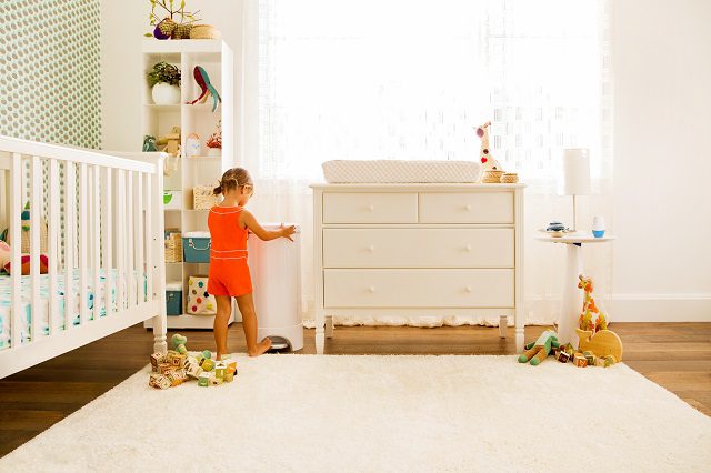 Toddler in orange outfit playing in a light wood nursery with white crib, dresser, and shelving unit filled with toys and books.