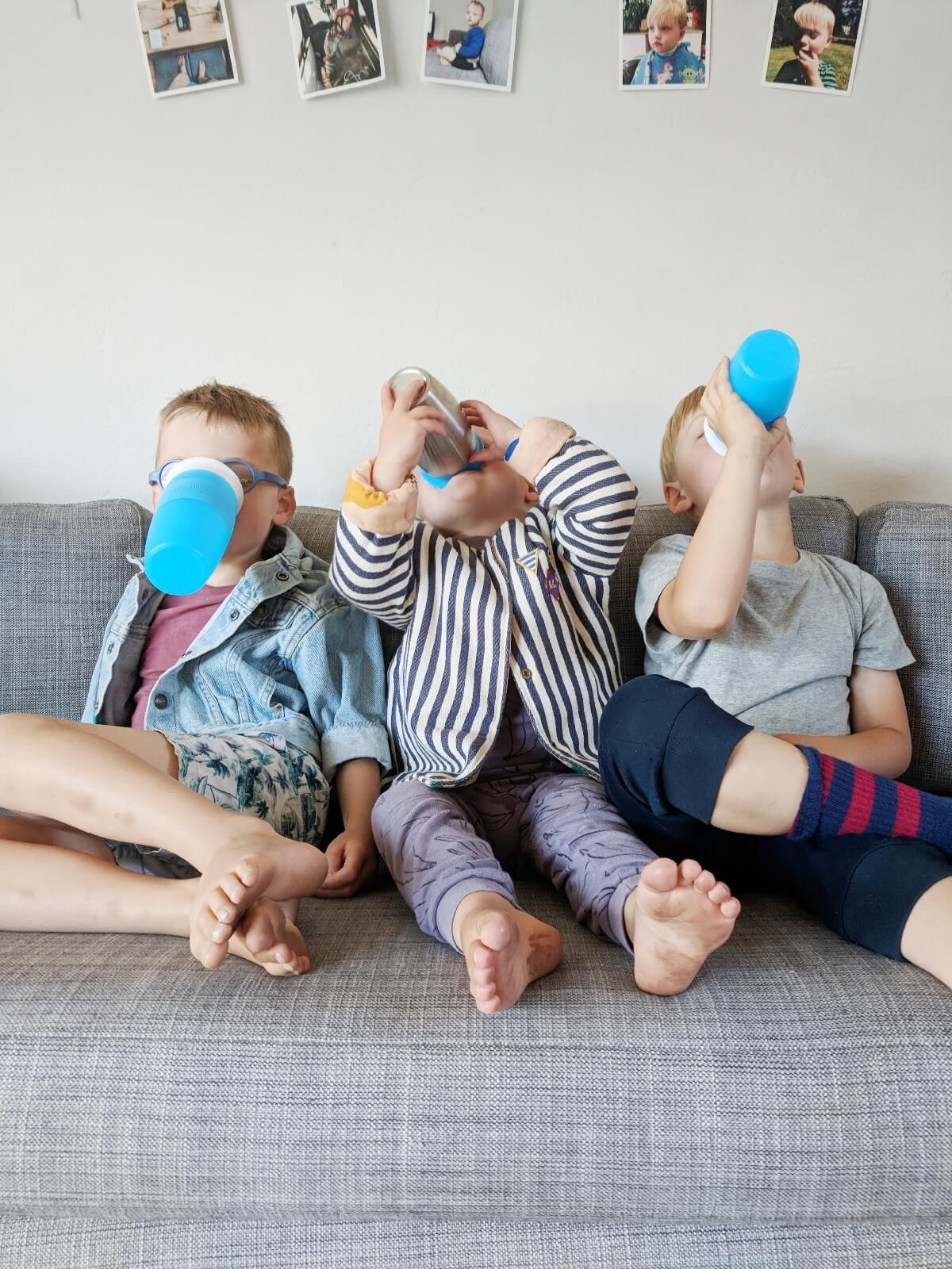 Three toddlers sitting on a couch drinking from blue Munchkin sippy cups with white straps, with family photos on the wall behind them.