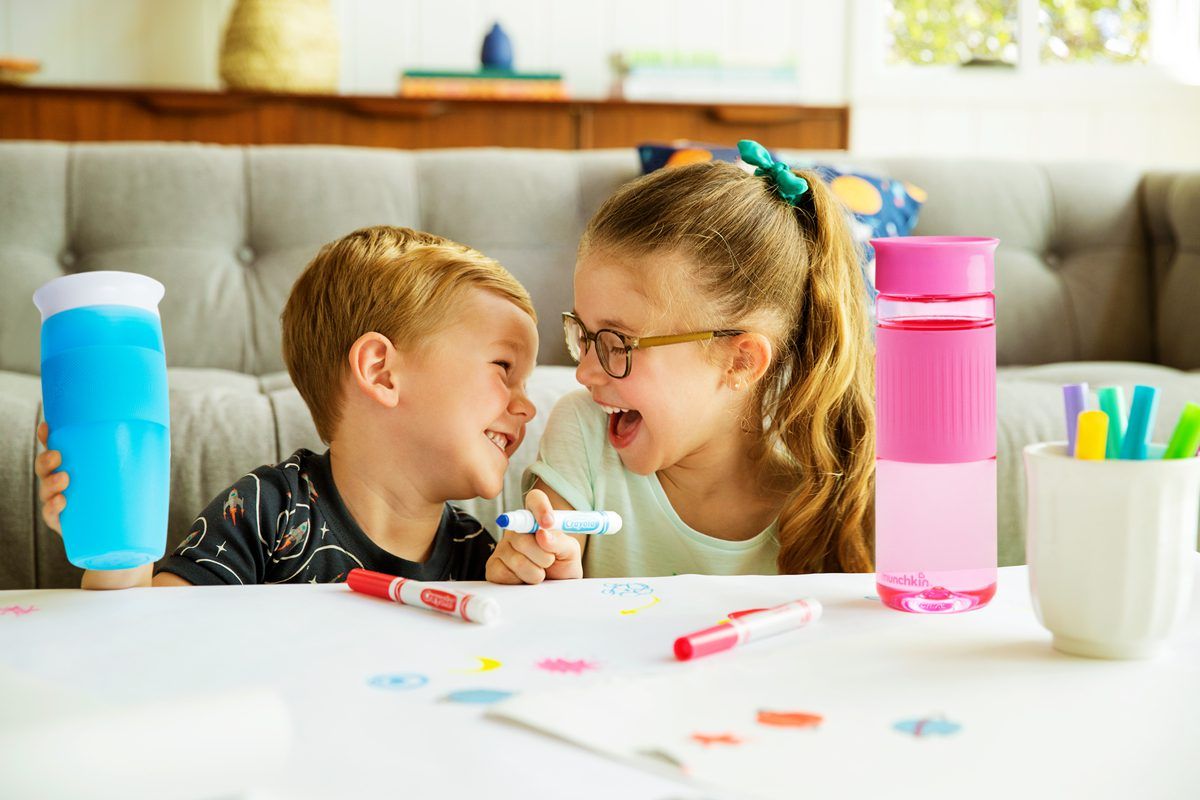Two children drawing and coloring together at a table with Munchkin sippy cups, markers, and art supplies nearby.