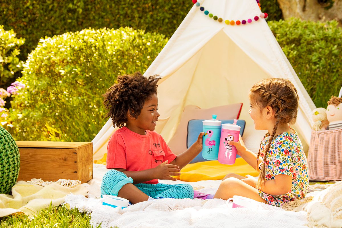 Two toddlers sitting in sand playing with Munchkin sippy cups inside a teepee tent in a garden setting with colorful decorations.