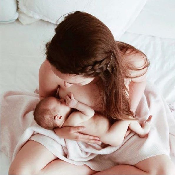 Mother kissing newborn baby while sitting on a white bed