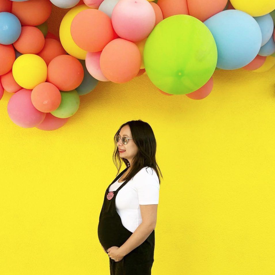 Pregnant woman wearing glasses and a black apron over a white shirt, smiling in front of a bright yellow wall decorated with colorful balloons in red, orange, pink, yellow, green, and blue.