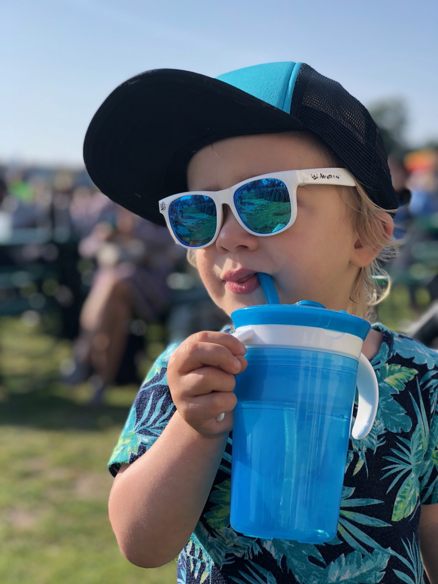 Toddler wearing a blue and black cap and white sunglasses, drinking from a blue Munchkin sippy cup outdoors at a crowded event.