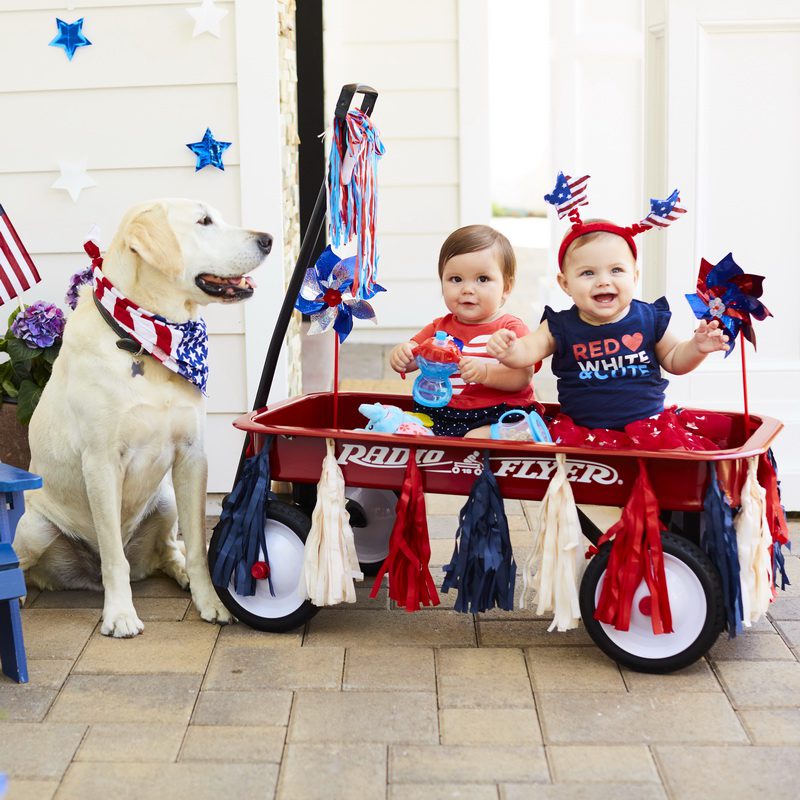 Two toddlers sitting in a red Radio Flyer wagon decorated with patriotic ribbons and flags, alongside a yellow Labrador Retriever wearing a stars and stripes bandana, with hanging stars and Fourth of July decorations in the background.