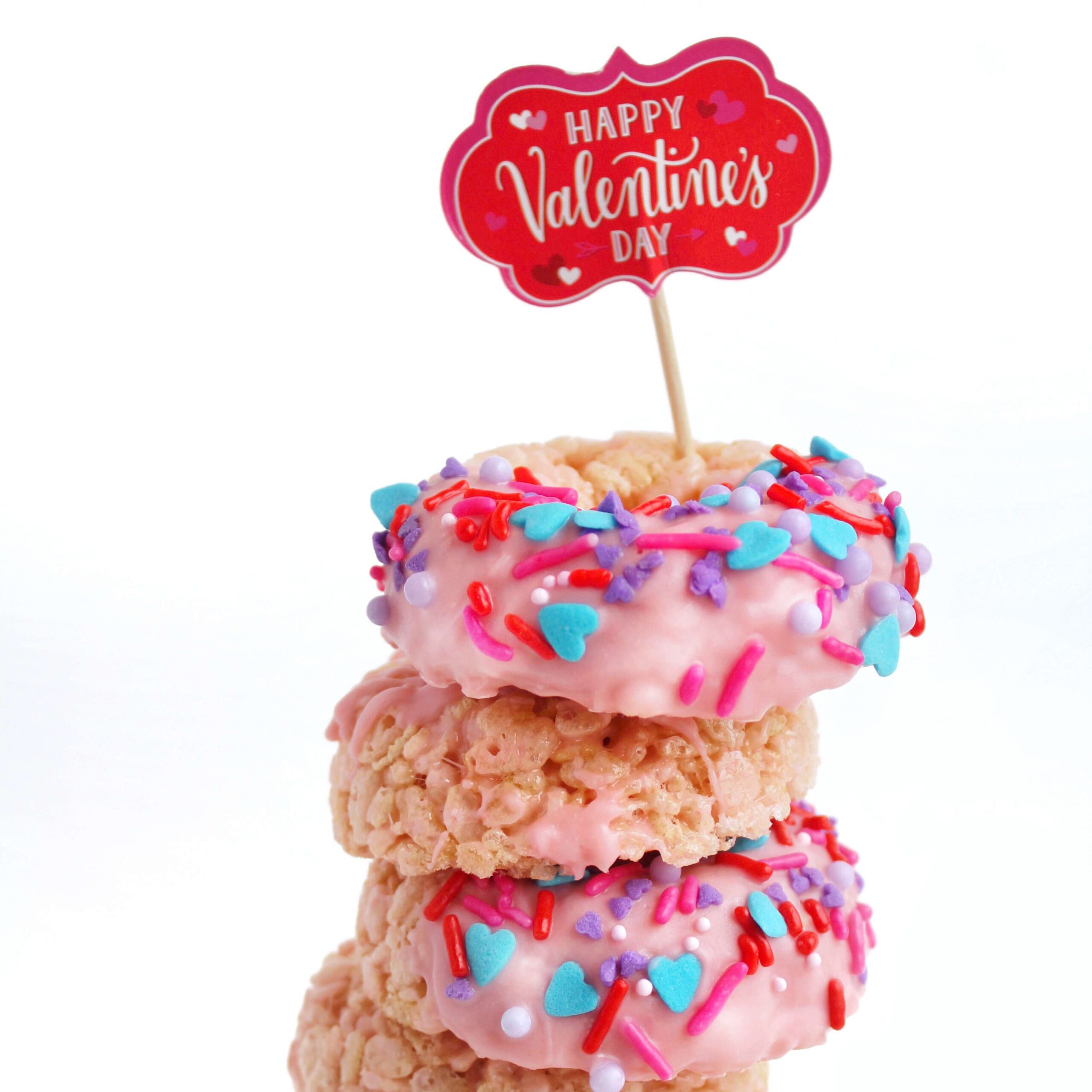 Stacked rice cereal treats with pink frosting, colorful heart and sprinkle decorations, and a Happy Valentine's Day cake topper.