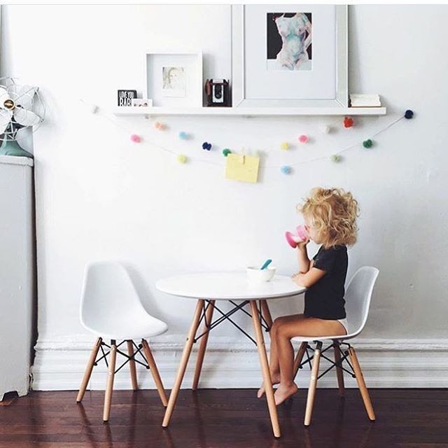 Toddler with curly hair sitting at a white round table with white chairs, drinking from a pink Munchkin cup in a bright playroom with shelving and colorful pom-pom garland on the wall.