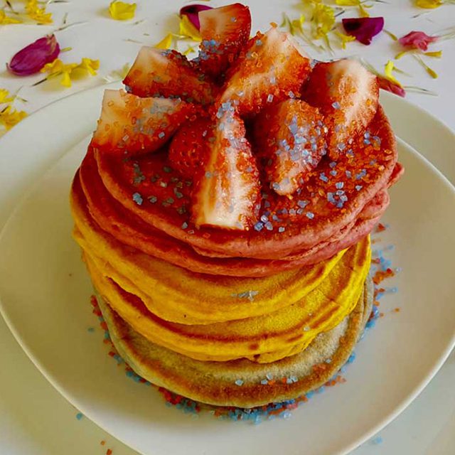 Colorful stacked pancakes with yellow and pink layers, topped with strawberry slices and sprinkles, on a white plate with flower petals.