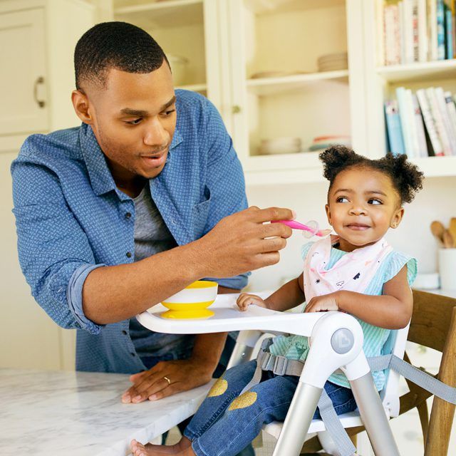 Man feeding toddler from a Munchkin high chair tray with a pink spoon and yellow bowl.