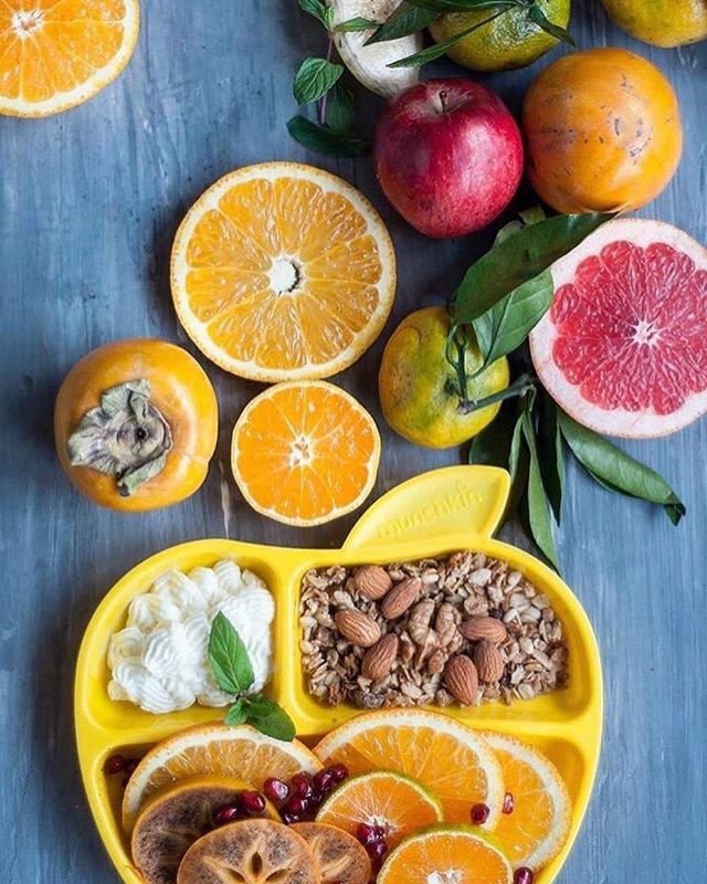 Yellow divided plate with fresh fruit and almonds, surrounded by oranges, grapefruit, apples, and citrus slices on blue surface.