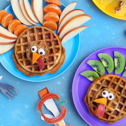 Creative breakfast food art featuring two waffles decorated as turkeys with fruit and vegetable feathers on blue and purple plates, arranged on a blue surface.