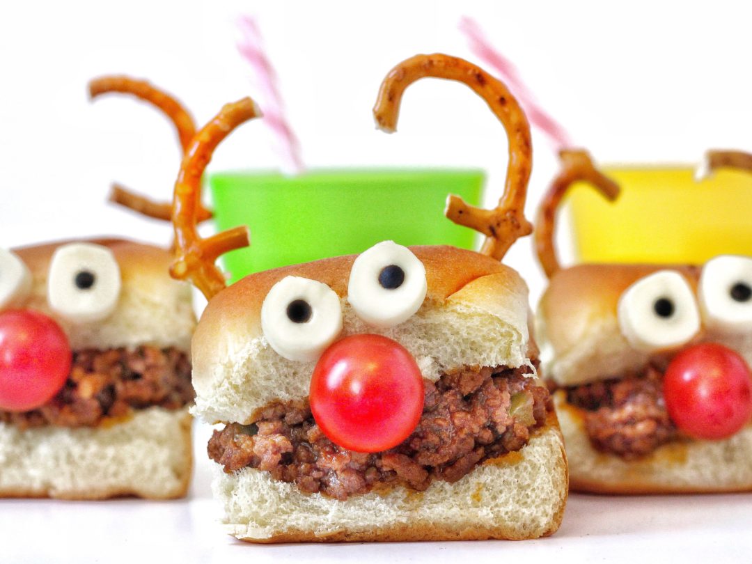 Reindeer-themed sandwiches made with bread, ground meat, white candy eyes, red gumball noses, and pretzel antlers, arranged on a white surface with colorful paper backgrounds.